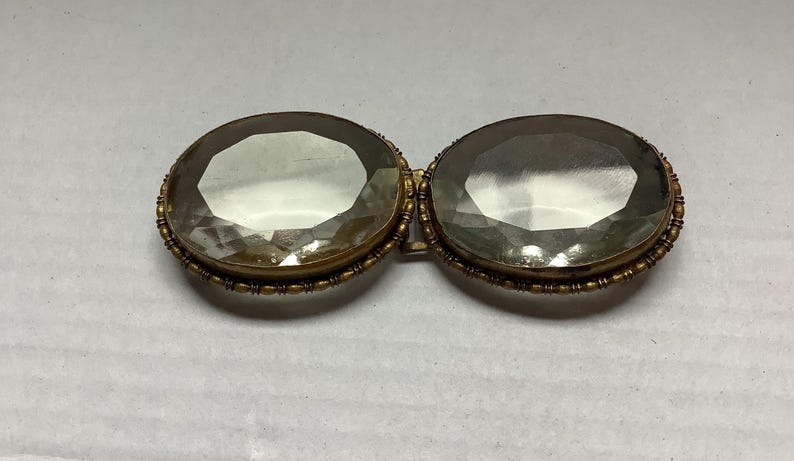 Pode incluir: Broche antigo com duas grandes pedras preciosas ovais e facetadas. As pedras est&atilde;o engastadas numa moldura de metal dourado com detalhes intrincados. Um acess&oacute;rio vintage, provavelmente da era vitoriana ou eduardiana.