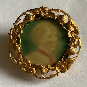 May include: A vintage gold-toned brooch with a portrait inside a floral frame. The portrait has a green background and features a person wearing a red tie. The brooch has a pin on the back.