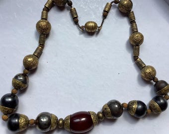 Pode incluir: Um colar vintage com uma variedade de contas em ouro, prata e preto. As contas apresentam padrões e texturas intrincados, com uma grande conta ovalada castanho-avermelhada no centro. O colar é enfiado em um cordão escuro e tem um fecho.