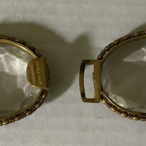 Pode incluir: Duas fivelas de cinto ovais douradas antigas com inser&ccedil;&otilde;es de vidro transparente facetado. As fivelas est&atilde;o abertas, mostrando o intrincado trabalho em metal e o vidro transparente. O metal tem um design detalhado de contas ao redor do vidro.