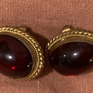 May include: A pair of gold-tone earrings with a dark red oval-shaped stone set in each. The stones are surrounded by a gold-tone rope-like design.