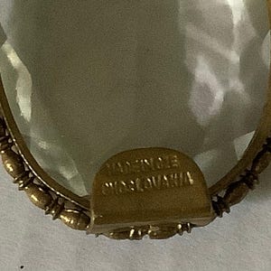 Pode incluir: Close-up de um pingente vintage em forma oval com um centro transparente e facetado. O pingente &eacute; emoldurado em metal dourado com uma borda decorativa de contas. A base do pingente tem o texto "MADE IN CZECHOSLOVAKIA".