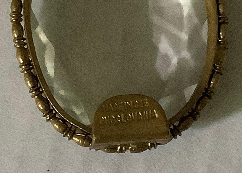 Pode incluir: Close-up de um pingente vintage em forma oval com um centro transparente e facetado. O pingente &eacute; emoldurado em metal dourado com uma borda decorativa de contas. A base do pingente tem o texto "MADE IN CZECHOSLOVAKIA".