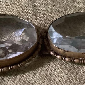 Pode incluir: Duas pedras ovais de vidro transparente em engastes de lat&atilde;o antigo com uma borda de contas. As pedras facetadas captam a luz. O fundo &eacute; um tecido texturizado de cor neutra.