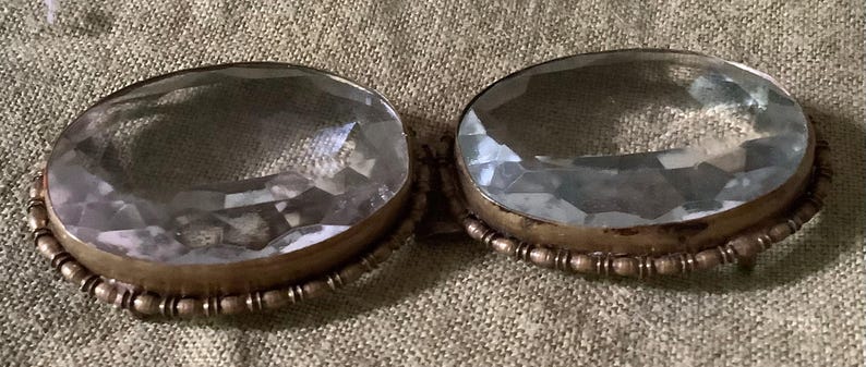 Pode incluir: Duas pedras ovais de vidro transparente em engastes de lat&atilde;o antigo com uma borda de contas. As pedras facetadas captam a luz. O fundo &eacute; um tecido texturizado de cor neutra.