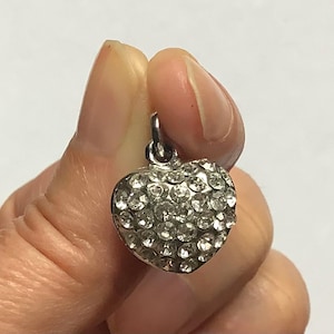 Op de afbeelding: Zilverkleurige hartvormige hanger, bedekt met kleine, heldere kristallen. De hanger heeft een kleine lus aan de bovenkant om aan een ketting of armband te bevestigen. Het hart wordt tussen vingers gehouden tegen een witte achtergrond.
