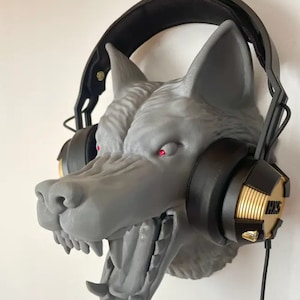 Könnte beinhalten: Graue Wolfskopfskulptur mit roten Augen, die schwarze und goldene Kopfhörer hält. Auf den Kopfhörern steht der Text "HKS". Die Skulptur ist an einer weißen Wand befestigt.