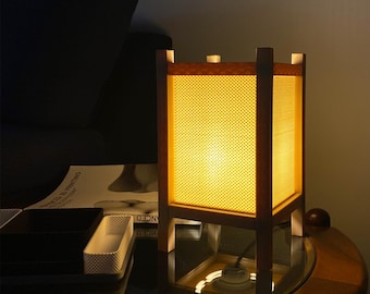 May include: A square table lamp with a wooden frame and a translucent, yellow, honeycomb-patterned shade. The lamp emits a warm, bright glow from within, illuminating the four vertical wooden posts that support the structure at each corner.