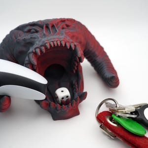 Könnte beinhalten: Ein rot-schwarzer 3D-gedruckter Monsterkopf mit offenem Maul, der einen weißen Würfel hält. Ein weißer Videospiel-Controller liegt auf dem Kopf des Monsters. Ein Schlüsselsatz mit einem schwarzen Schlüsselanhänger befindet sich im Vordergrund.