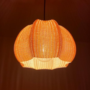 Op de afbeelding: Een oranje hanglamp met een geweven textuur. De lamp heeft een afgeronde vorm met een geschulpte bovenkant en een warme gloed die van binnenuit komt. Hij hangt aan een zwart snoer tegen een donkere achtergrond.