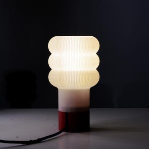 Op de afbeelding: Een moderne tafellamp met een uniek ontwerp. De lamp heeft een gestapelde, afgeronde kap met verticale lijnen, die een zacht, warm licht uitstraalt. De basis loopt over van wit naar rood, met een effen rode onderkant. Een zwart netsnoer loopt uit de basis.