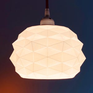 Pode incluir: Uma luminária pendente branca com uma superfície geométrica facetada e um padrão triangular repetitivo. A cúpula da lâmpada é iluminada por dentro, emitindo um brilho suave, e está suspensa por um fio escuro preso a um soquete branco.