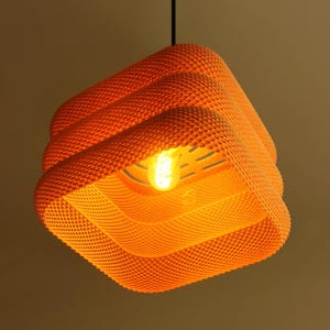 Op de afbeelding: Oranje, vierkante hanglamp met een getextureerd, gestippeld oppervlak. De lamp heeft een gelaagd ontwerp en een zichtbare gloeilamp in het midden. Hij hangt aan een zwart snoer.