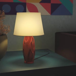 Könnte beinhalten: Eine moderne Tischlampe mit cremefarbenem Lampenschirm und korallenfarbenem Sockel mit offenem, fließendem Design. Die Lampe steht auf einer türkisfarbenen Oberfläche, mit geometrischen Kunstwerken im Hintergrund. Links befindet sich eine dekorative Vase.