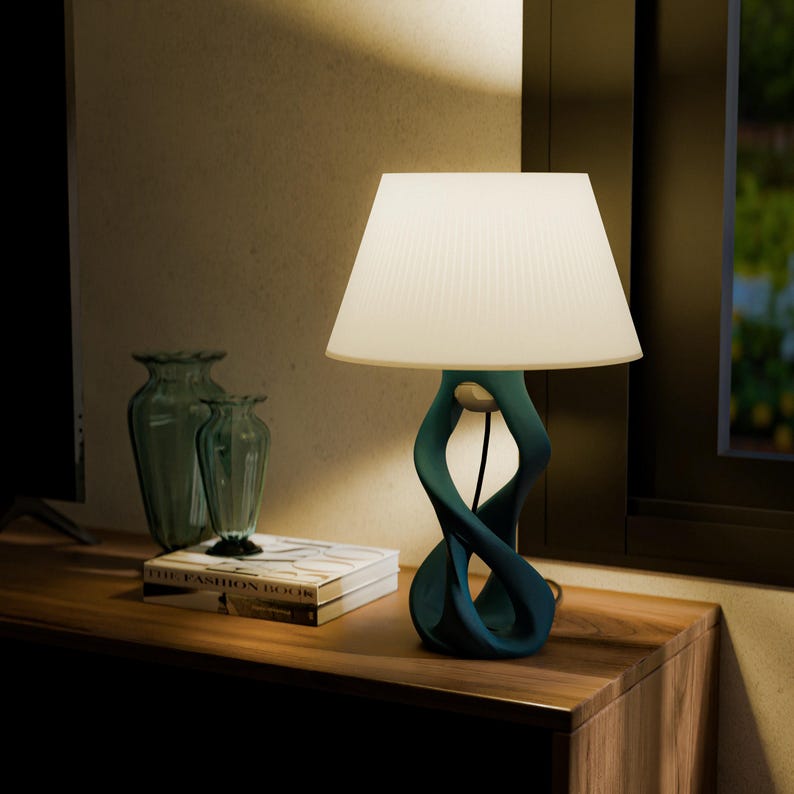 May include: A teal-colored table lamp with a white pleated lampshade. The base has a unique, intertwined design. The lamp sits on a wooden surface, next to two glass vases and a book titled "THE FASHION BOOK".