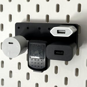 Może przedstawiać: Czarny uchwyt ścienny z czterema otworami trzyma trzy różne rodzaje ładowarek do telefonu. Ładowarki są białe, czarne i szare. Uchwyt ścienny jest przymocowany do białej tablicy z otworami.