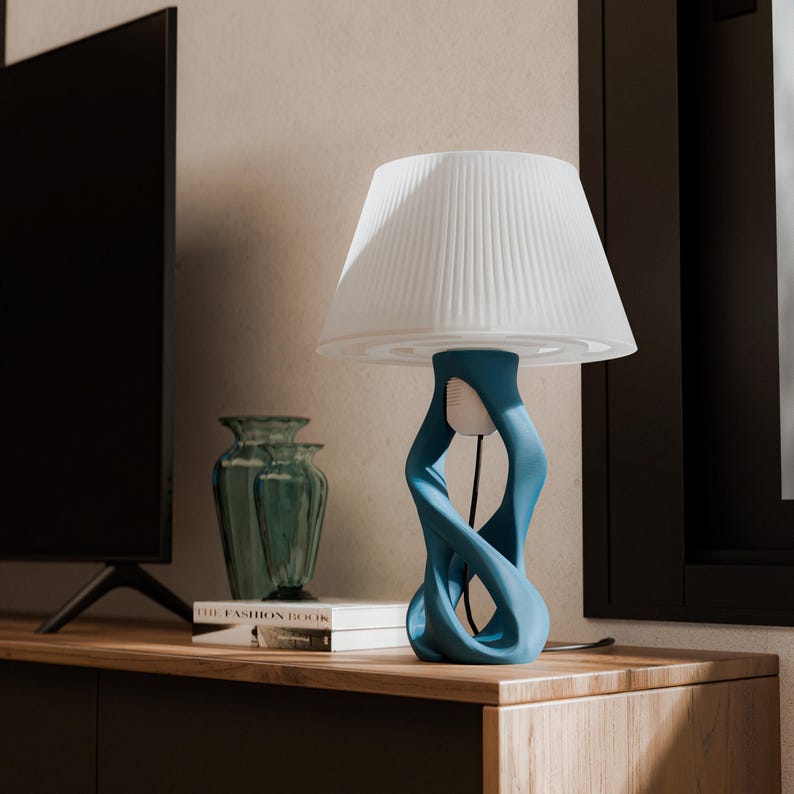 May include: A modern table lamp with a white pleated lampshade and a teal blue, abstract base. The lamp sits on a wooden surface, next to a teal vase and a book. The lamp's design is contemporary and artistic.