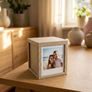 Könnte beinhalten: Ein quadratischer Holzbilderrahmen mit beigefarbener Oberfläche und gerippter Struktur an den Kanten. Der Rahmen enthält ein quadratisches Foto eines lächelnden Paares am Strand. Der Rahmen hat ein schlichtes und klares Design.