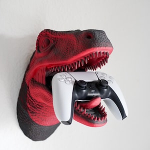 Peut inclure: Un support mural en forme de tête de dinosaure rouge et noir avec une manette de jeu vidéo blanche et noire dans sa gueule.