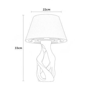 May include: A white table lamp with a pleated lampshade, 22 cm wide and 33 cm tall. The lamp base has an abstract, intertwined design. The lamp is a line drawing.