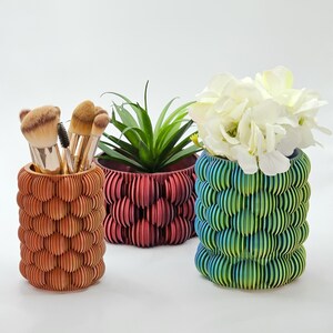 Könnte beinhalten: Drei dekorative Töpfe mit einem gewellten Design. Der Topf links ist braun, der mittlere Topf ist rot und der Topf rechts ist grün. Der mittlere Topf hat eine kleine grüne Pflanze darin und der rechte Topf hat weiße Blumen darin.