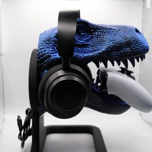 Może przedstawiać: Stojak na słuchawki w kształcie niebieskiego dinozaura z czarnymi słuchawkami i białym kontrolerem w paszczy.