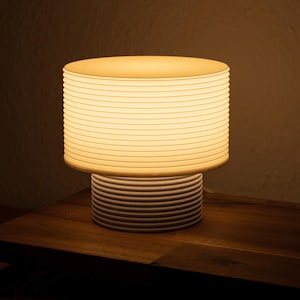 Puede incluir: Una lámpara de mesa con una base cilíndrica y una pantalla cilíndrica más ancha a juego, ambas construidas con una serie de anillos horizontales apilados. La lámpara emite un brillo cálido y suave que ilumina la textura acanalada del material blanco roto.