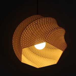 Op de afbeelding: Een unieke, geometrische hanglamp met een textuur, geweven patroon in een warme, goudbruine kleur. De lamp heeft een opvallend, open-bodem ontwerp, met een heldere, witte gloeilamp. De armatuur hangt aan een donker snoer tegen een zwarte achtergrond.