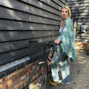 May include: A light blue crocheted shawl with a floral pattern. The shawl is worn over a white top and blue jeans. A bicycle with a black basket is in the foreground. The background features a dark gray wooden wall and a brick wall.