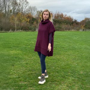 May include: A burgundy poncho with a turtleneck collar and ribbed detailing at the hem and sleeves. The poncho is worn over a striped long-sleeve shirt and dark blue leggings. The person is wearing gray sneakers on a grassy field.