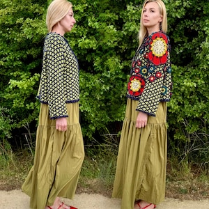 May include: Two women wearing olive green maxi dresses and red shoes stand in front of a green leafy background. One woman wears a navy and yellow crochet jacket, the other a colorful floral crochet jacket.