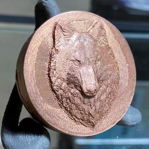 May include: A close-up of a copper-colored, round, 3D printed pendant featuring a detailed wolf head design.