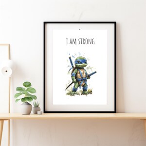 May include: Framed art print with a watercolour illustration of a cartoon turtle character wearing a blue mask and holding two katanas. The text "I AM STRONG" is printed above the character. The artwork is displayed in a black frame.