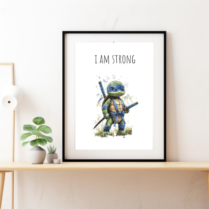 May include: Framed art print with a watercolour illustration of a cartoon turtle character wearing a blue mask and holding two katanas. The text "I AM STRONG" is printed above the character. The artwork is displayed in a black frame.