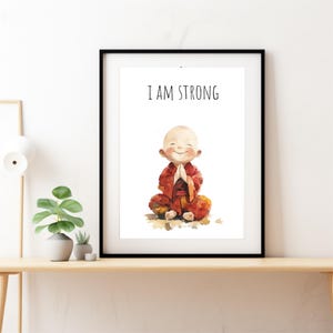 May include: A framed print with a watercolor illustration of a smiling cartoon monk in a red robe sitting in a meditative pose. The text "I AM STRONG" is printed in black above the illustration.