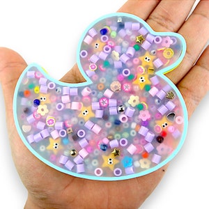 Puede incluir: Una bandeja de pato en forma de pato de resina transparente azul claro llena de cuentas de colores, estrellas y otros pequeños encantos. La bandeja está sentada en la mano de una persona.