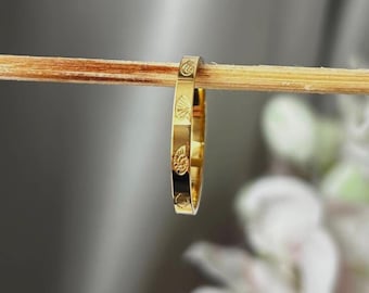 Peut inclure: Une bague dorée ornée de motifs de coquillages gravés. La bague est présentée sur un bâton en bois, mettant en valeur son design. La surface de la bague reflète la lumière, soulignant les motifs de coquillages. Il s'agit d'un bijou.