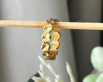 Peut inclure: Une bague dorée avec un motif répétitif de découpes circulaires et en forme d'éclat d'étoile. La bague est présentée sur une cheville en bois sur un fond flou avec des accents floraux blancs et verts. Le design de la bague est symétrique et détaillé.