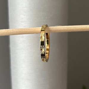 May include: A gold ring featuring a repeating starburst design. The thin ring is made of metal. It is displayed on a wooden dowel against a white backdrop. The ring's design is etched into the metal.