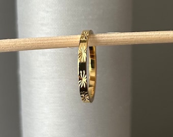 Peut inclure: Une bague dorée ornée d'un motif d'éclats d'étoiles répétés. La bague fine semble être en métal. Elle est présentée sur une cheville en bois sur fond blanc. Le motif est gravé.