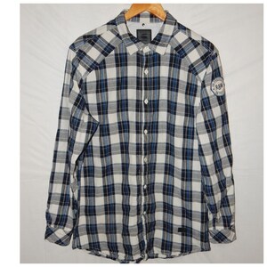 Könnte beinhalten: Ein langärmeliges Button-Down-Hemd in einem Karomuster aus Weiß, Marineblau und Grau. Das Hemd hat einen Kragen und einen kleinen Aufnäher am oberen linken Ärmel mit dem Text "RAW". Das Hemd hängt an einem Holzbügel.