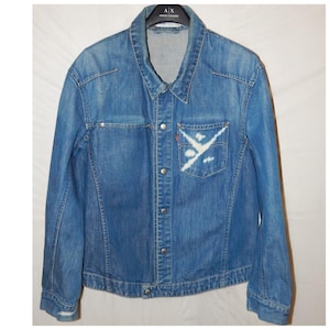 Puede incluir: Chaqueta vaquera azul con cierre de botones, cuello y mangas largas. La chaqueta tiene un bolsillo con un diseño blanco y una etiqueta roja. La chaqueta es de mezclilla de lavado medio.