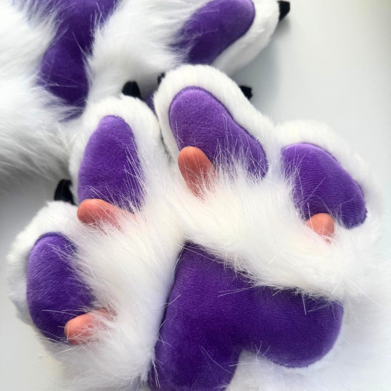 May include: Close-up of a pair of furry paw gloves. The gloves feature white fur with purple paw pads and black claws. The pads have a rounded shape, and the claws are small and pointed.