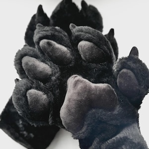 May include: A pair of black plush animal paw gloves featuring soft faux fur, dark grey padded paw pads, and pointed black claws. The gloves are designed with a textured, fuzzy exterior and distinct toe sections, mimicking the appearance of a large animal paw with a prominent central palm pad.