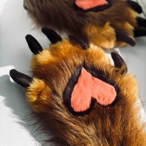May include: A pair of brown faux fur paw slippers with black claws and a coral heart-shaped accent on the paw pads. The slippers are designed to resemble animal paws.