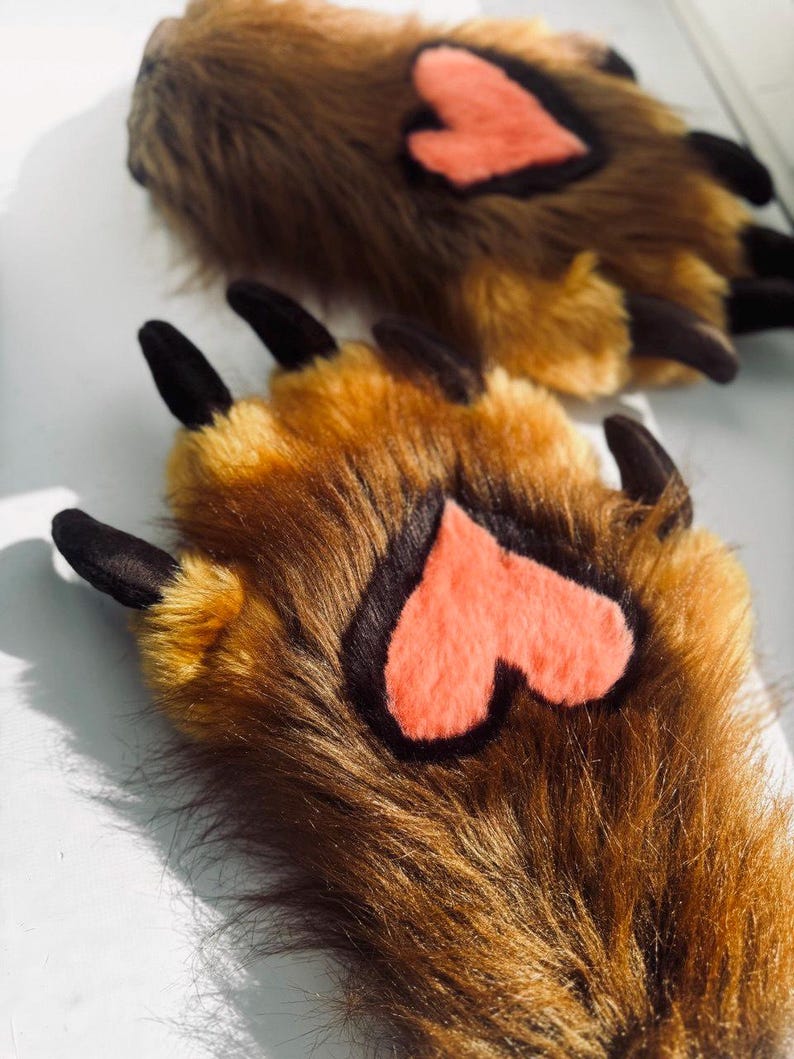 May include: A pair of brown faux fur paw slippers with black claws and a coral heart-shaped accent on the paw pads. The slippers are designed to resemble animal paws.