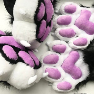 May include: Close-up of plush paws in white, black, and purple. The paws have white fur with black accents and purple paw pads. The fur is soft and fluffy, and the overall design is cute and playful.