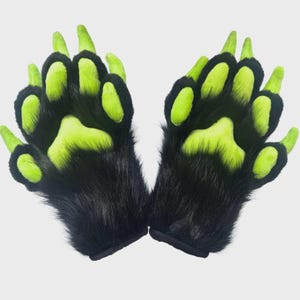 May include: A pair of plush gloves designed as animal paws, featuring long black faux fur covering the back and wrist areas, contrasted with bright neon green paw pads and claw-like tips on each of the five fingers for a textured appearance.