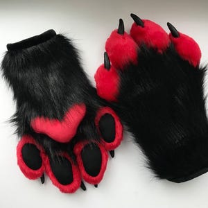 May include: A pair of fingerless gloves designed as animal paws, featuring long black faux fur, bright red plush paw pads, and pointed black claws. The gloves have a soft, textured appearance with distinct padded sections for the fingers and palm.