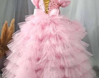 May include: A pink tulle dress with a gold sequined bodice and a large bow at the waist. The dress has multiple layers of tulle and is perfect for a special occasion.